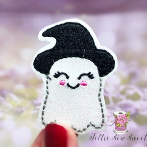 May include: A white ghost with a black witch hat and pink cheeks. The ghost is smiling and has eyelashes. The ghost is embroidered on a white fabric.