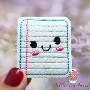 May include: A white, blue, and pink embroidered patch of a sheet of lined paper with a smiling face. The paper has a glittery texture.