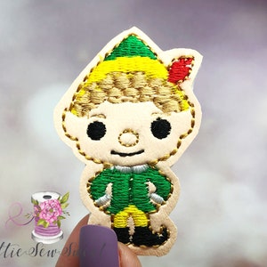 May include: A felt appliqué of a cartoon elf with a green hat, yellow and red accents, and a green and yellow outfit. The elf has a smiling face with black eyes and a gold nose.