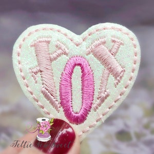 May include: A white heart-shaped patch with pink embroidered letters spelling "XO" on a white glitter background.