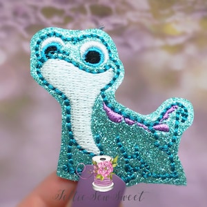 May include: A teal glittery embroidered applique in the shape of a cartoon lizard. The applique has a white belly, black eyes, and purple accents. The edges are outlined with teal stitching. The logo "Jollie Sew Sweet" is in the bottom right corner.