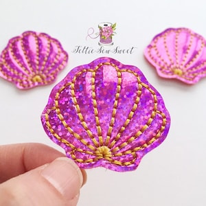 May include: Three seashell-shaped appliques in shades of pink and purple with gold stitching. The central applique is held by a hand. The appliques have a glittery texture. The text "Feltie Sew Sweet" is in the upper right.