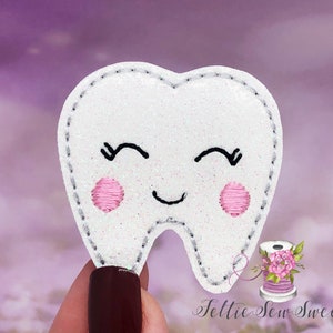 May include: A white felt tooth with a smiling face, pink cheeks, and black eyelashes. The tooth is decorated with glitter and has a gray stitched outline.