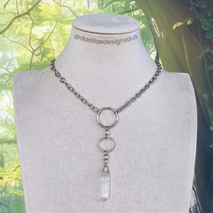 Può includere: Una collana color argento con catena e tre ciondoli circolari. Una punta di cristallo trasparente pende dal ciondolo inferiore. La collana è esposta su un manichino. Il testo "@darkfaedesignsau" è visibile.