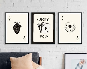 Puede incluir: Tres impresiones enmarcadas con fondo blanco e ilustraciones negras. Las impresiones muestran un corazón, una mano de cartas de juego y un corazón con una corona. El texto en la impresión del medio dice "LUCKY YOU."