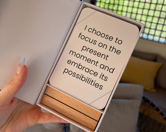 Puede incluir: Una caja rectangular blanca que contiene una baraja de cartas y dos bloques de madera de color marrón claro. La tarjeta superior presenta el texto impreso en negro: I choose to focus on the present moment and embrace its possibilities.