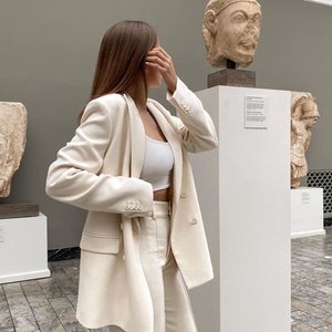 May include: A woman wearing a white pantsuit in a museum setting. She is standing next to a white pedestal with a sculpture on top. The sculpture is a bust of a person with a crown.