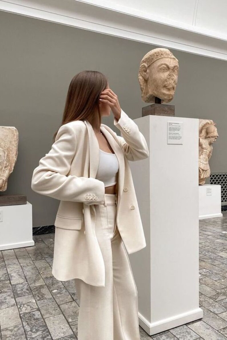 May include: A woman wearing a white pantsuit in a museum setting. She is standing next to a white pedestal with a sculpture on top. The sculpture is a bust of a person with a crown.