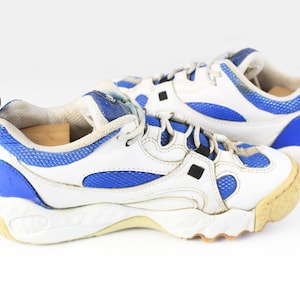 Może przedstawiać: Para białych i niebieskich but&oacute;w sportowych z grubą podeszwą. Buty mają niebieski panel z siatki i białą sk&oacute;rzaną cholewkę.