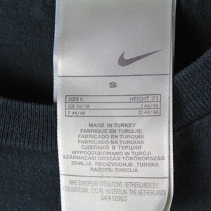 Peut inclure: &Eacute;tiquette blanche avec du texte noir indiquant la taille S, GB 36/38, F 44/48, Hauteur 173, 144/48, D 44/48. L'&eacute;tiquette indique &eacute;galement "Fabriqu&eacute; en Turquie" dans plusieurs langues. L'&eacute;tiquette est attach&eacute;e &agrave; un t-shirt noir.