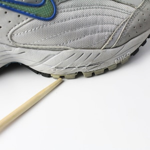 May include: A close-up of the bottom of a gray and white running shoe with a blue and green swoosh logo. The shoe has a black rubber outsole with a tread pattern. The shoe is sitting on a white surface.