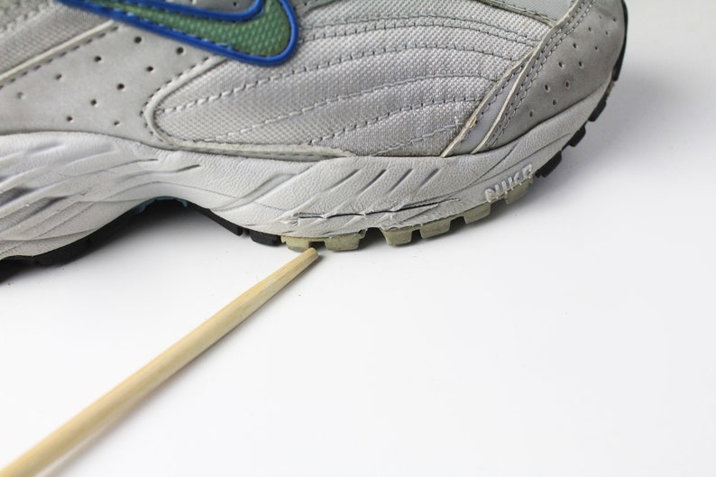 May include: A close-up of the bottom of a gray and white running shoe with a blue and green swoosh logo. The shoe has a black rubber outsole with a tread pattern. The shoe is sitting on a white surface.