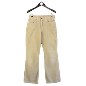 Può includere: Un paio di pantaloni in velluto a coste color marrone chiaro appesi a un appendiabiti in legno. I pantaloni hanno una gamba larga e una vestibilità dritta.