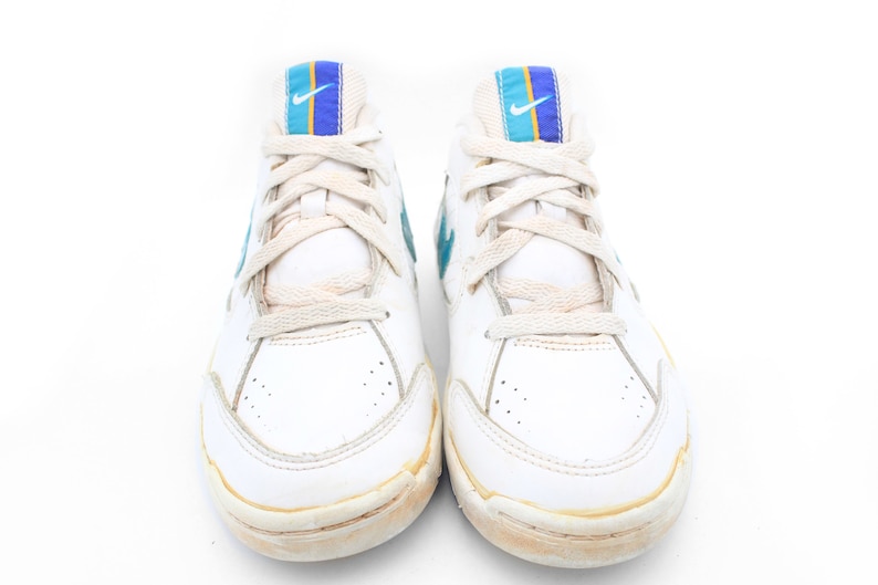 May include: White leather sneakers with blue and yellow accents on the tongue. The laces are white and the shoes have a worn look.