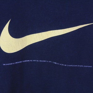 Peut inclure: Un sweat-shirt bleu marine avec un grand logo Nike swoosh jaune. Le texte "THE SWOOSH IS A REGISTERED TRADEMARK NO. 977190. DESIGNED IN 1971. CURRENTLY WORN BY CHAMPIONSHIP ATHLETES, EVERYWHERE." est imprim&eacute; sous le logo.