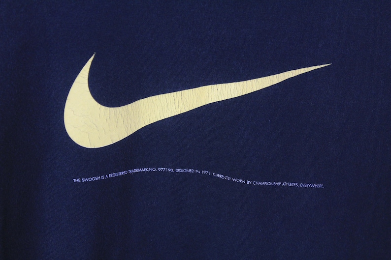 Peut inclure: Un sweat-shirt bleu marine avec un grand logo Nike swoosh jaune. Le texte "THE SWOOSH IS A REGISTERED TRADEMARK NO. 977190. DESIGNED IN 1971. CURRENTLY WORN BY CHAMPIONSHIP ATHLETES, EVERYWHERE." est imprim&eacute; sous le logo.