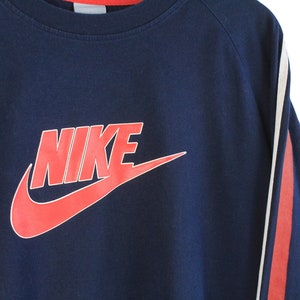 May include: A navy blue sweatshirt with red and white stripes on the sleeves. The sweatshirt features a large red Nike logo with the word "NIKE" in white.