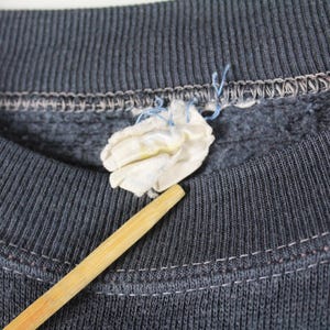 May include: Close-up of the neckline of a dark gray sweatshirt with a small white tag attached. The sweatshirt has a ribbed texture and a sewn-in seam around the neckline.