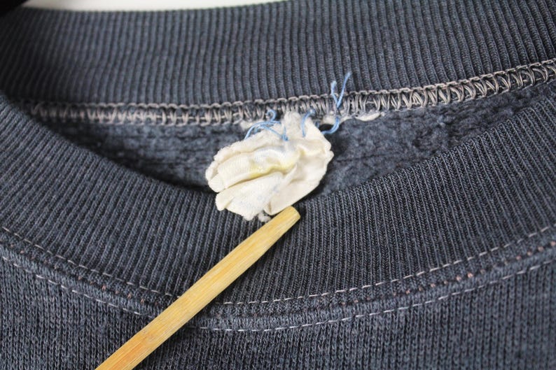 May include: Close-up of the neckline of a dark gray sweatshirt with a small white tag attached. The sweatshirt has a ribbed texture and a sewn-in seam around the neckline.