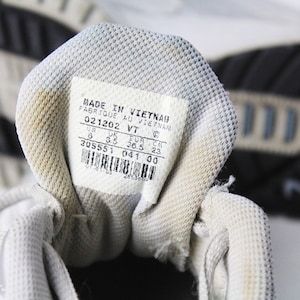 May include: Close-up of the inside of a white athletic shoe with a white label that reads "MADE IN VIETNAM FABRIQUE AU VIETNAM 021202 VT 6 3.5 36.5 23 EUR CM 305551 041 00".