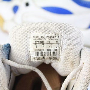 Może przedstawiać: Zbliżenie wnętrza białego buta sportowego z niebieskimi akcentami. Etykieta rozmiaru buta zawiera napis "MADE IN INDONESIA", "970204 IA", "US 8", "UK 7", "EUR 41", "CH 26", "144031 101 00".