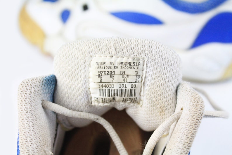 Może przedstawiać: Zbliżenie wnętrza białego buta sportowego z niebieskimi akcentami. Etykieta rozmiaru buta zawiera napis "MADE IN INDONESIA", "970204 IA", "US 8", "UK 7", "EUR 41", "CH 26", "144031 101 00".