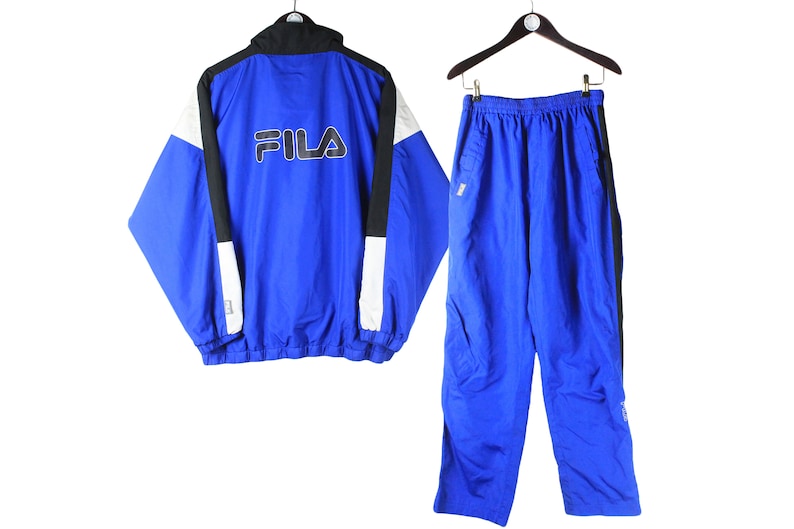 May include: A blue Fila tracksuit with black and white accents. The jacket has a zipper closure and a Fila logo on the front. The pants have a drawstring waist and side pockets.