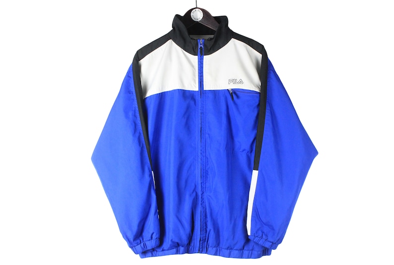 May include: A blue, white, and black Fila windbreaker jacket with a zipper closure. The jacket has a white panel across the chest with the Fila logo in black.