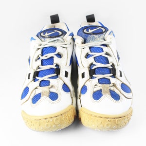 Może przedstawiać: Para białych but&oacute;w sportowych z niebieskimi akcentami i czarnym logo Swoosh. Buty mają białą siateczkową cholewkę i białą gumową podeszwę z fakturowanym wzorem.