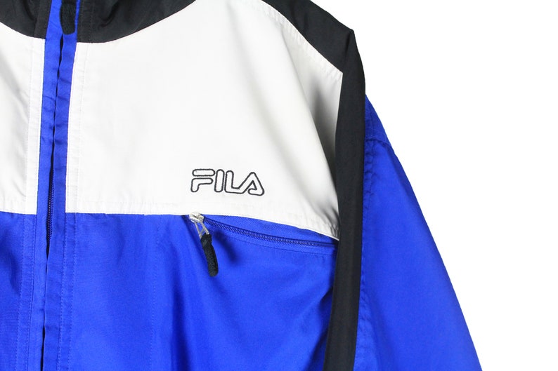 May include: A blue, white, and black windbreaker jacket with a zipper closure and the word "FILA" printed on the white panel.