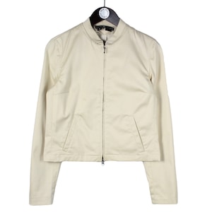 Puede incluir: Una chaqueta de color crema con cremallera, mangas largas y dos bolsillos.