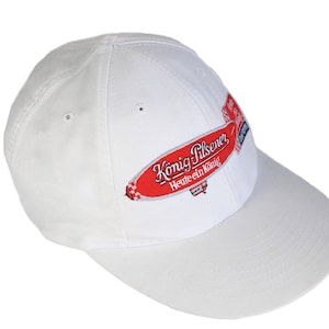 May include: White baseball cap with a red and white embroidered patch that says "König Pilsener Heute ein König".