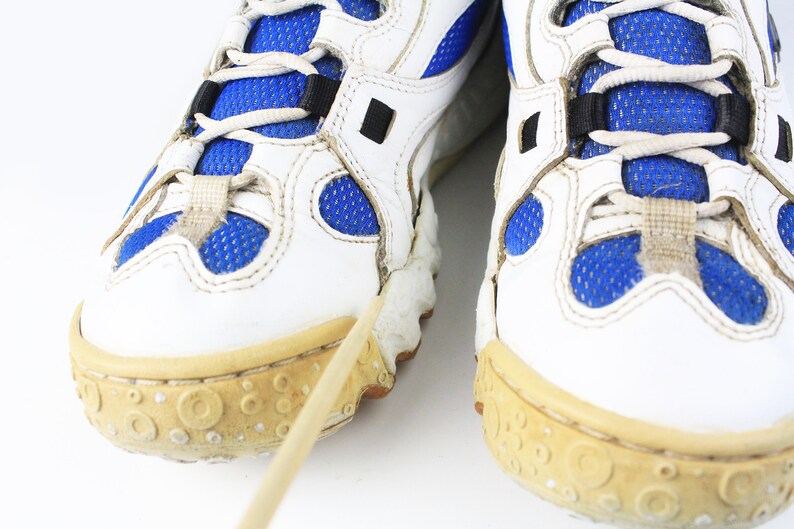 Może przedstawiać: Para białych but&oacute;w sportowych z niebieskimi wstawkami z siateczki i białymi sznurowadłami. Buty mają grubą, fakturowaną podeszwę.