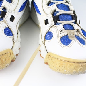 Może przedstawiać: Para białych but&oacute;w sportowych z niebieskimi siatkowymi wstawkami i brązowymi gumowymi podeszwami. Buty mają czarne sznur&oacute;wki i czarny pasek na g&oacute;rze.