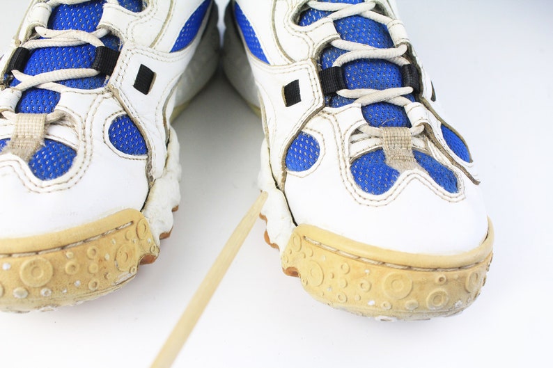 Może przedstawiać: Para białych but&oacute;w sportowych z niebieskimi siatkowymi wstawkami i brązowymi gumowymi podeszwami. Buty mają czarne sznur&oacute;wki i czarny pasek na g&oacute;rze.