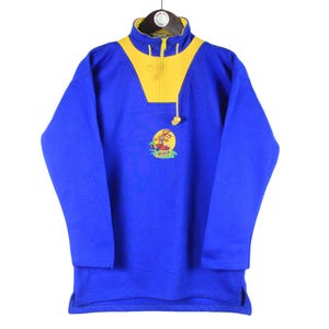 Puede incluir: Un jersey de forro polar azul con cuello y detalles en los hombros amarillos. El jersey tiene un pequeño diseño bordado en el pecho. La parte superior tiene una cremallera de cuarto y un cordón. La prenda está colgada en una percha.