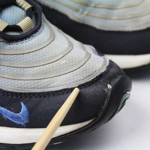 May include: Close-up of a pair of blue and black Nike Air Max 97 sneakers. The shoes have a mesh and leather upper with a white midsole and black outsole. The Nike swoosh logo is visible on the side of the shoe.