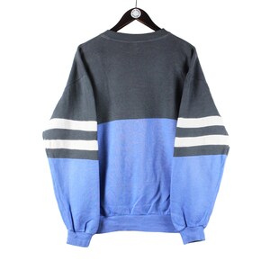 May include: A blue and gray sweatshirt with white stripes on the sleeves. The sweatshirt has a crew neck and a relaxed fit.