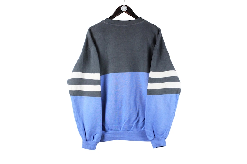 May include: A blue and gray sweatshirt with white stripes on the sleeves. The sweatshirt has a crew neck and a relaxed fit.