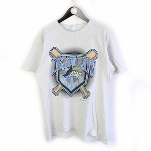Puede incluir: Una camiseta gris con un logotipo vintage de los Tampa Bay Devil Rays. El logotipo presenta un escudo azul y blanco con un pez raya diablo y las palabras "Devil Rays" en una fuente azul y blanca. El logotipo está rodeado por dos bates de béisbol cruzados.