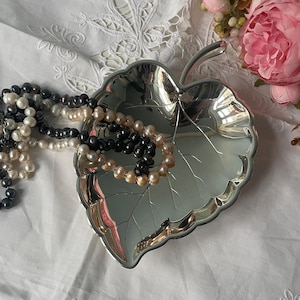 Può includere: Un piatto d'argento a forma di foglia contiene fili di perle nere, bianche e color pesca. Il piatto è appoggiato su una superficie bianca ricamata, con un bouquet di fiori rosa sullo sfondo. Le perle sono drappeggiate sul piatto.