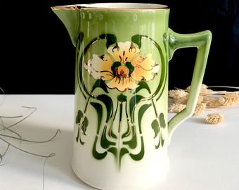 May include: A vintage ceramic jug with a green and white floral design. The jug features a yellow flower in the centre, surrounded by green leaves and gold accents. The handle and spout are also green, with a gold trim. A decorative piece for the home.