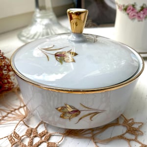 Könnte beinhalten: Eine weiße Porzellan-Zuckerdose mit Deckel, Goldrand und einem goldenen Griff. Die Schale ist mit einem floralen Design in Gold, Rot und Grün verziert. Die Schale steht auf einer braunen Spitzendecke.