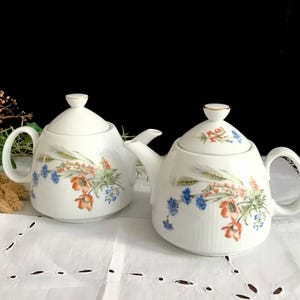 Könnte beinhalten: Zwei weiße Keramik-Teekannen mit Deckeln, Griffen und Ausgüssen. Jede Teekanne ist mit einem Blumendesign mit orangefarbenen, blauen und grünen Blumen und Weizenähren verziert. Die Teekannen stehen auf einer weißen Tischdecke.