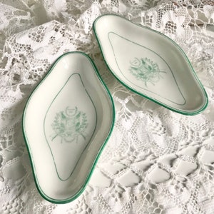 Può includere: Due piatti da portata in ceramica bianca con bordo verde e un motivo floreale verde. I piatti hanno la forma di un ovale allungato con un'estremità appuntita. Il design include uno stemma con il testo "GENT 1818".