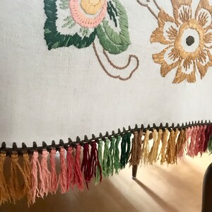 May include: Close-up of a white textile with embroidered floral designs in pink, yellow, and green. The bottom edge features a row of colorful tassels in shades of pink, green, and tan, adding a decorative touch to the fabric.