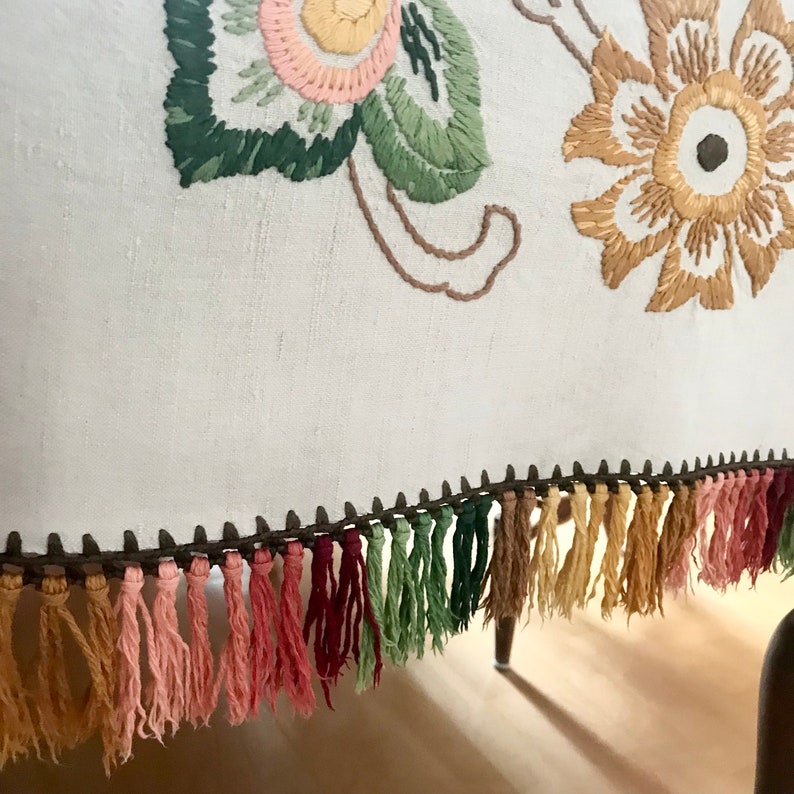 May include: Close-up of a white textile with embroidered floral designs in pink, yellow, and green. The bottom edge features a row of colorful tassels in shades of pink, green, and tan, adding a decorative touch to the fabric.