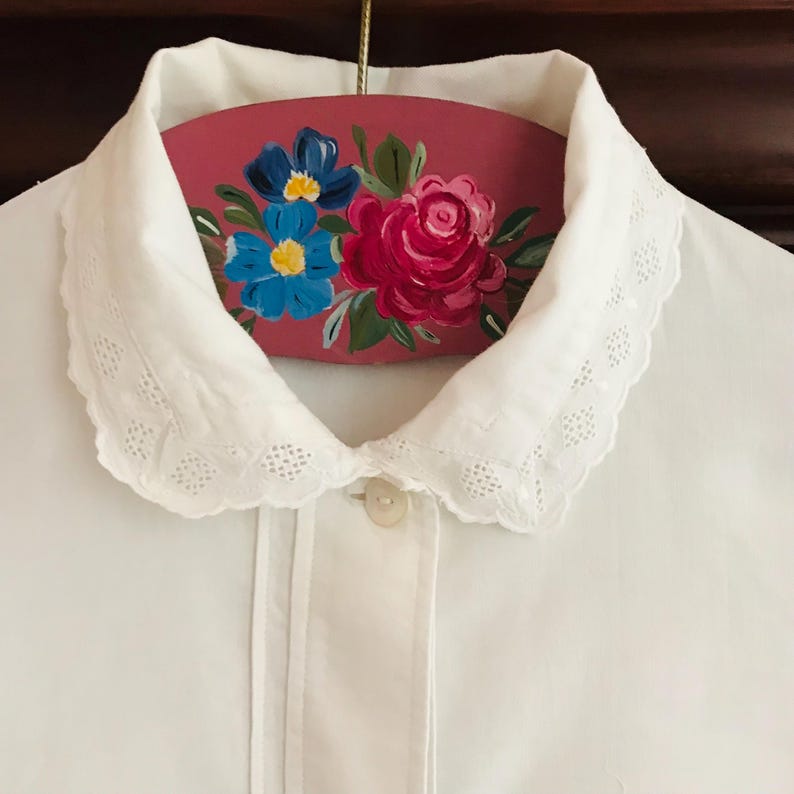 May include: White cotton blouse with a lace trim collar. The collar is folded over and has a pink hanger with painted flowers on it.