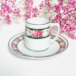 Può includere: Servizio da tè bianco con un motivo floreale a rose. La tazza ha una fascia nera con rose rosa e foglie verdi. Il piattino ha un bordo floreale abbinato. Il set è su una superficie bianca.