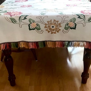 May include: White tablecloth with embroidered floral design in pink, green, and gold. The edges are trimmed with a black border and colorful tassels. The tablecloth is draped over a wooden table.